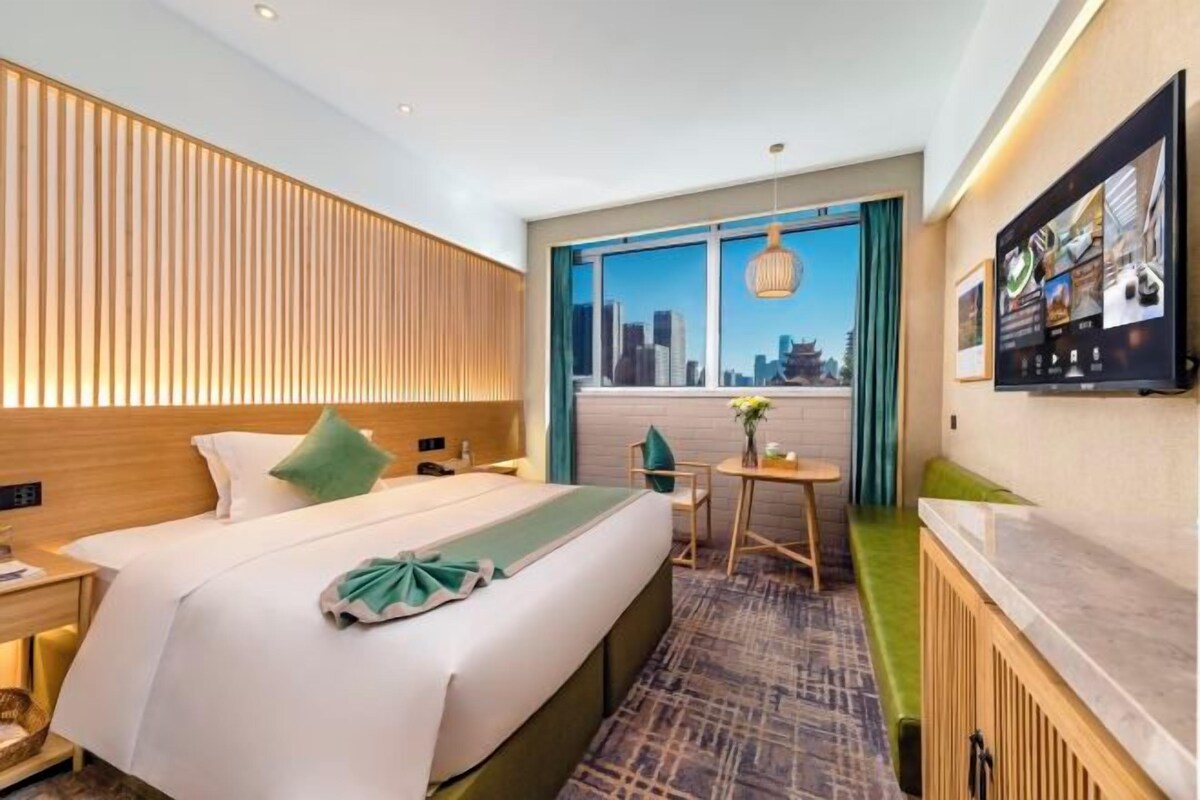 A hotel room with a bed, a desk, a chair, a window with a city view, and a wall-mounted TV.