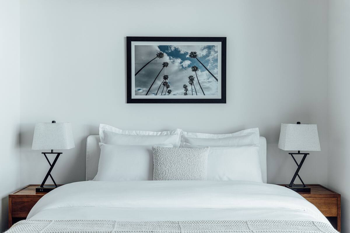 A neatly made bed with white linens, two bedside tables with lamps, and a framed picture of palm trees on the wall.