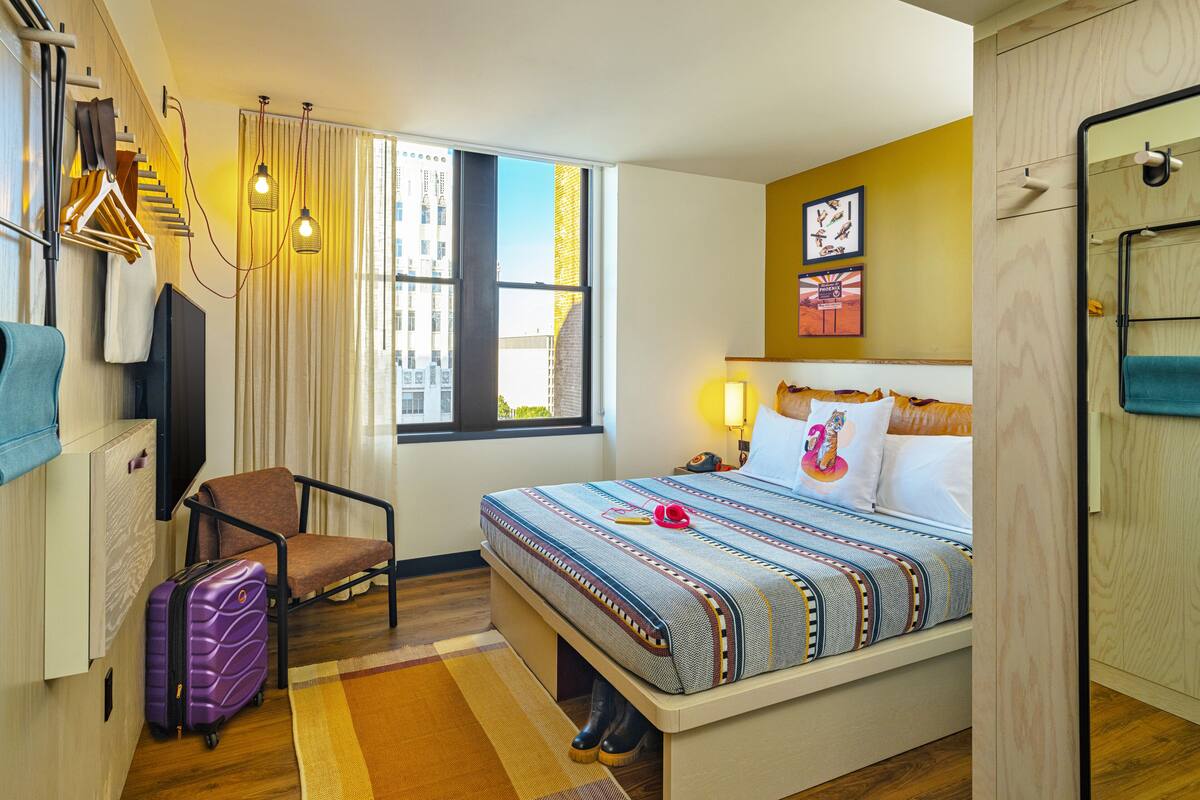 Ein Hotelzimmer mit Bett, Stuhl, Koffer, Fernseher und Fenster mit Blick auf die Stadt.