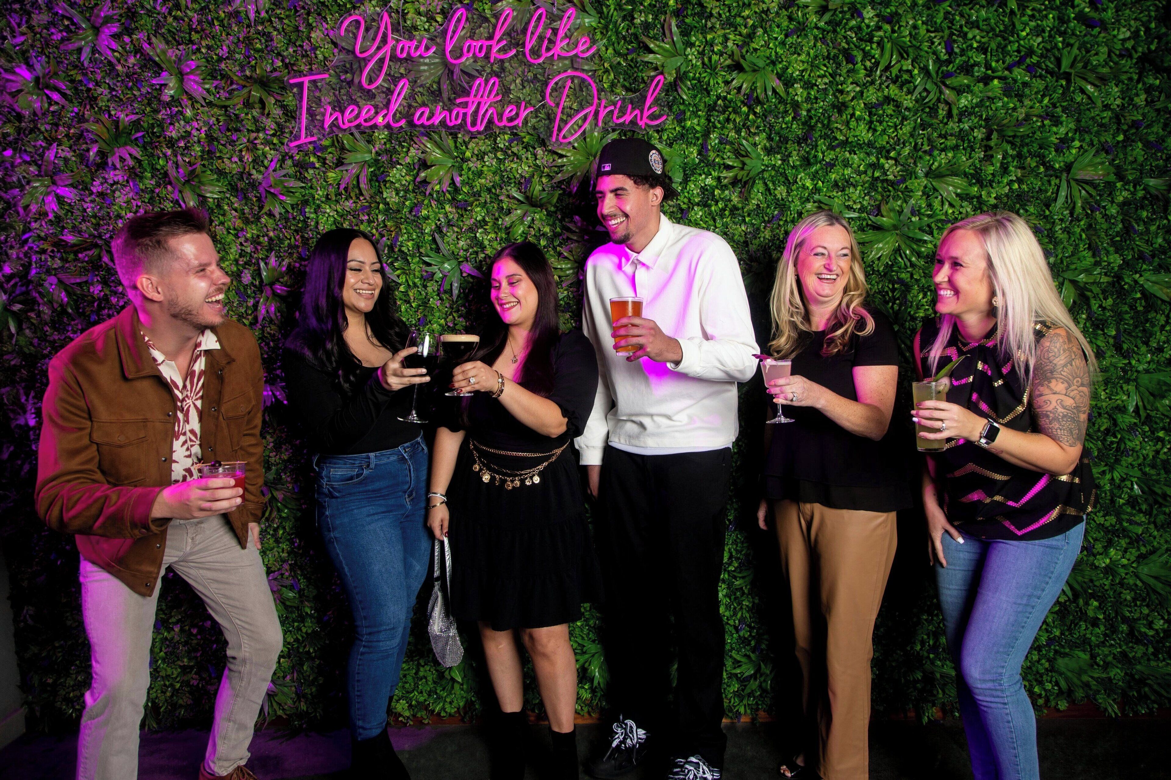 A group of people standing in front of a green leafy wall with a neon sign that reads "You look like I need another drink."