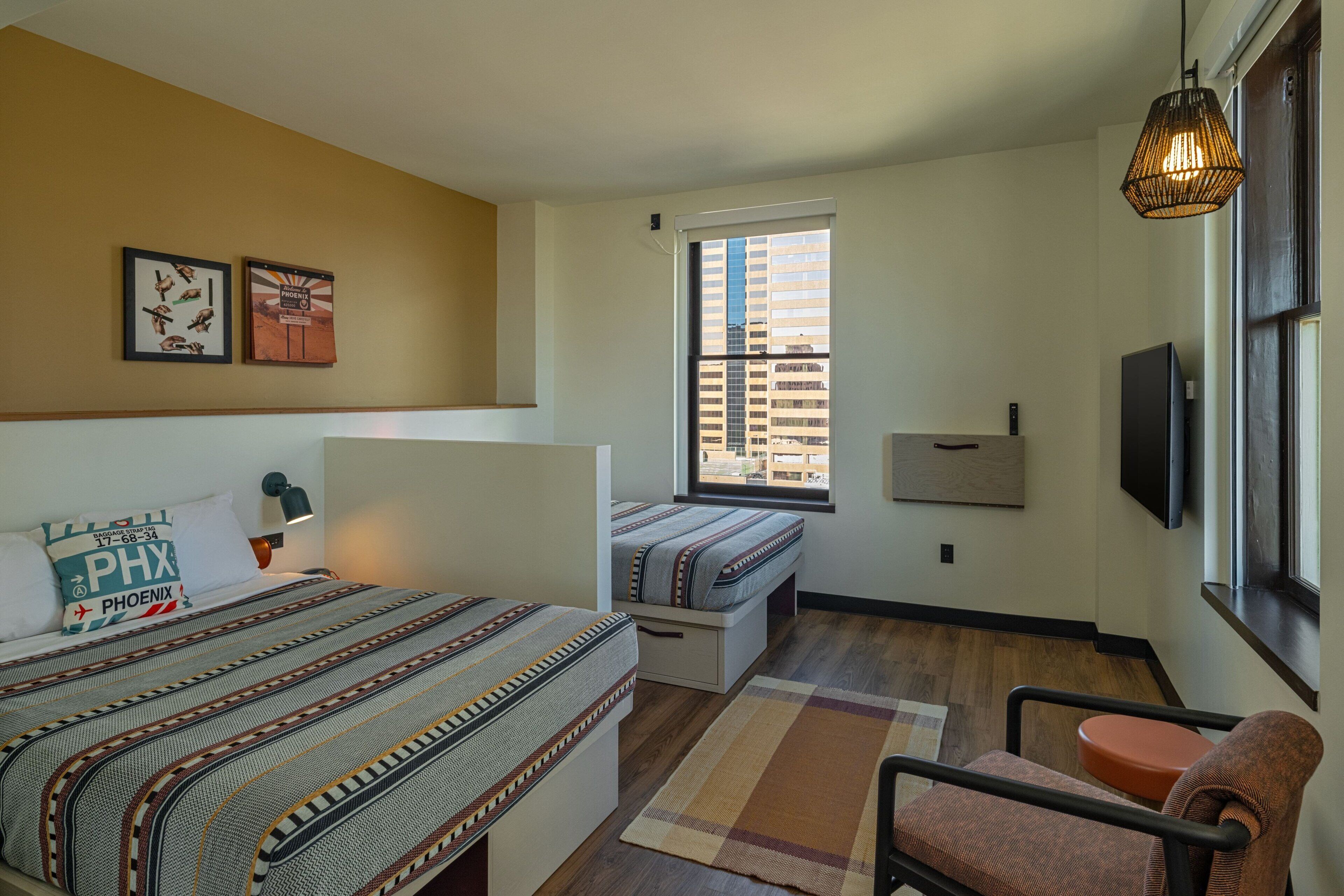 Quarto de hotel com cama, escrivaninha, cadeira, janela com persianas e televisão.