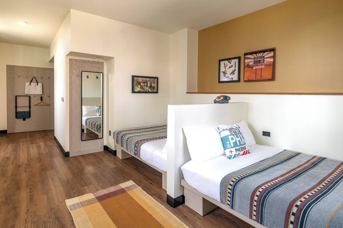 Ein Hotelzimmer mit zwei Einzelbetten, Holzboden, Spiegel, kleinem Schreibtisch und einem Wandtelefon.