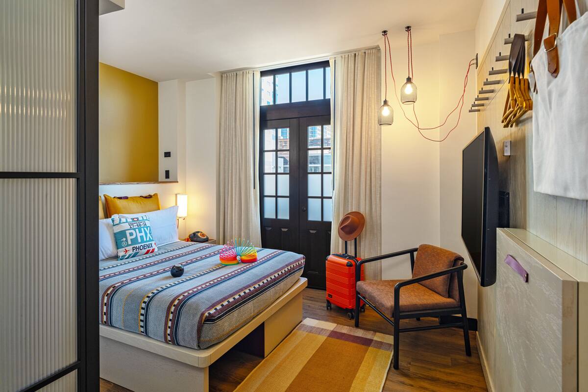 Ein Hotelzimmer mit Bett, Stuhl, Koffer, Fernseher und einem Fenster mit Vorhängen.