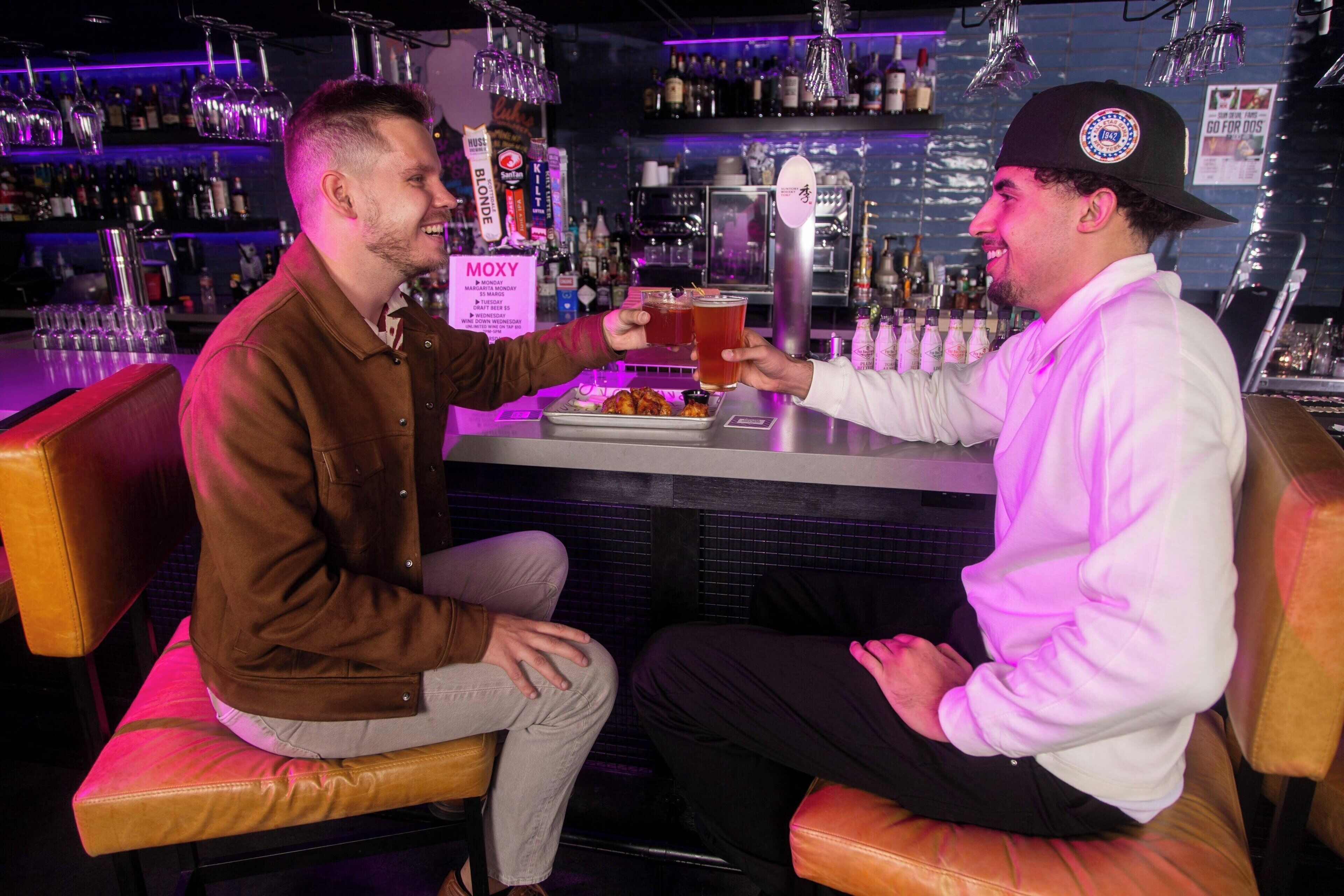 Two people seated at a bar with a drink in each hand, a menu sign, and various bottles and glasses on shelves in the background.