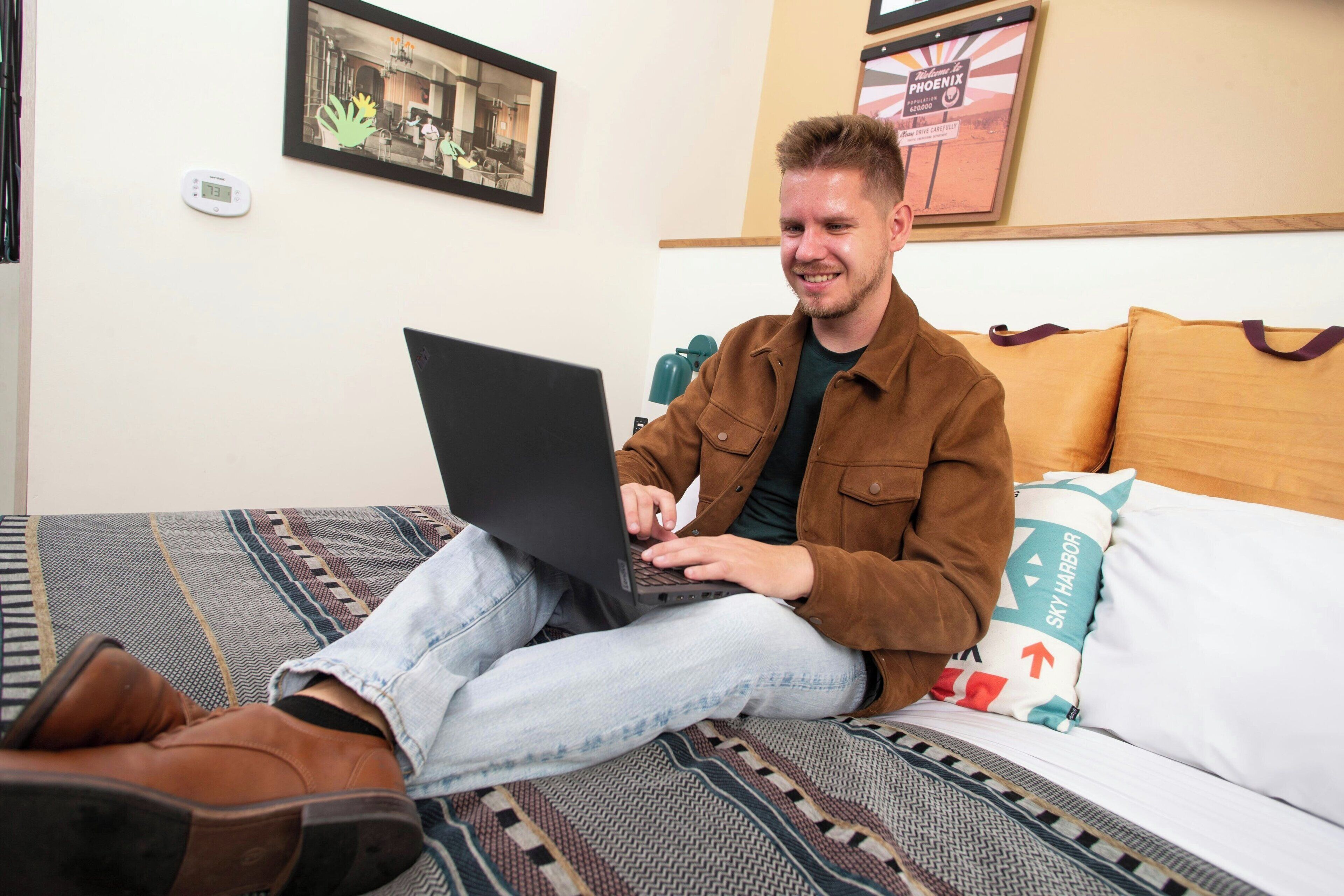 A person sitting on a bed with a laptop, a framed photo on the wall, and a sign with "Welcome to Phoenix" text.