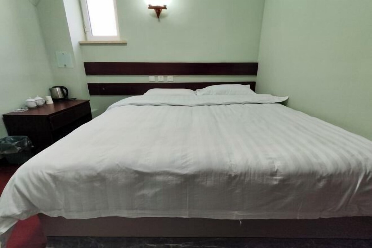 Habitación individual con una cama que tiene cabecera de madera, una mesita de noche con hervidor y una ventana con lámpara.