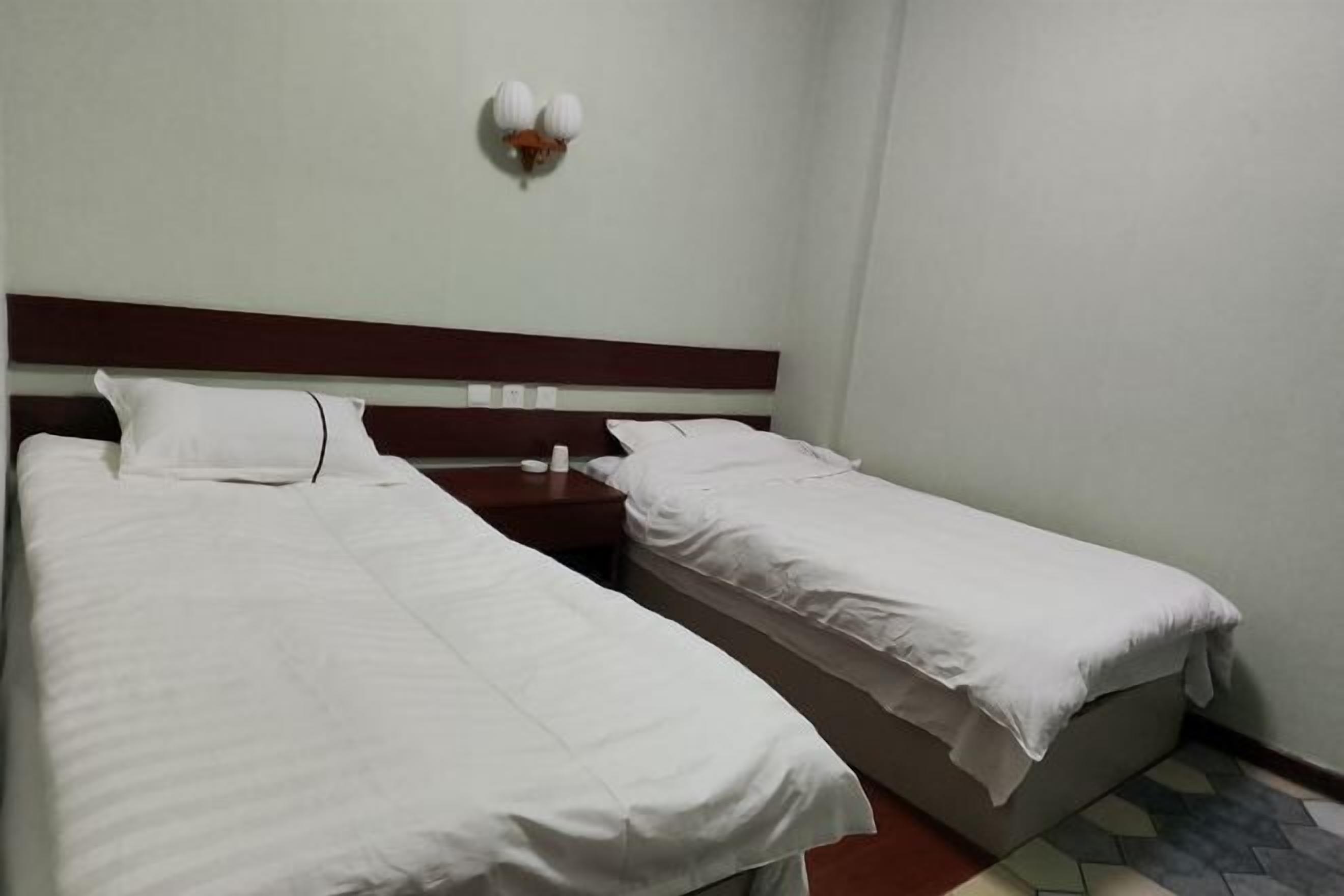 A room with two single beds, a wooden headboard, a bedside table with a lamp, and a tiled floor.