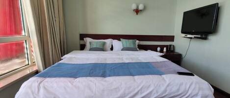 Quarto de hotel com cama, televisão fixada na parede e janela com cortinas.