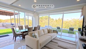 A modern living room with a large beige sofa, a glass dining table, and chairs. There are large windows offering a view of a garden and trees.