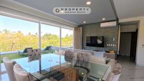 A modern living room with a glass-top dining table surrounded by chairs, a flat-screen TV mounted on the wall, and a view of the outdoors.