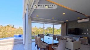 A modern living room with a glass dining table, comfortable seating, a flat-screen TV, and a view of greenery outside.