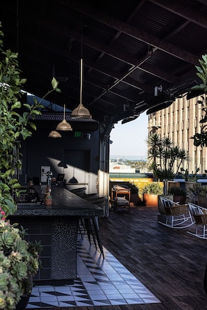 An outdoor bar area with a wooden floor, hanging pendant lights, and potted plants. - The Aster (Los Angeles)