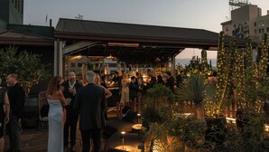 Un área de patio al aire libre con mobiliario, plantas y una sección cubierta donde personas están cenando.