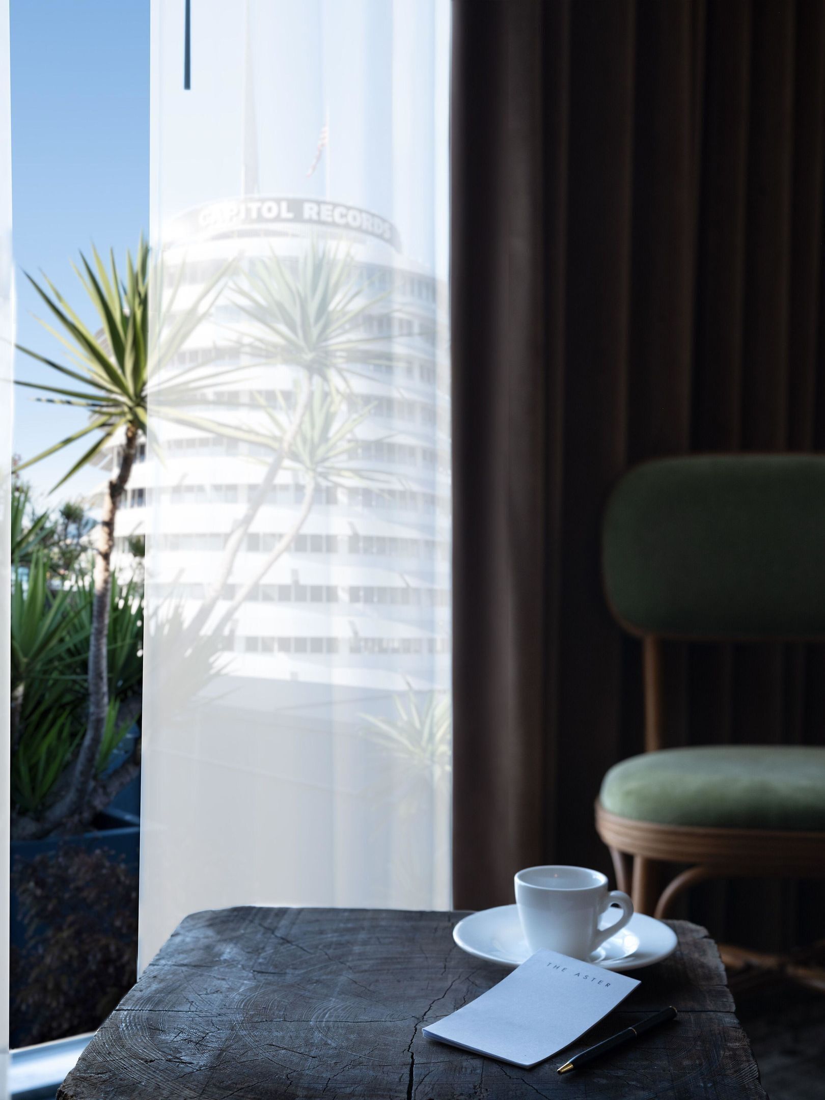 A table with a cup, a notepad, and a pen, a chair, and a view of a building through a window.