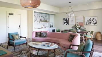 A living room with a pink sofa, teal chairs, a round coffee table, and a dining area with a wooden table and chairs.