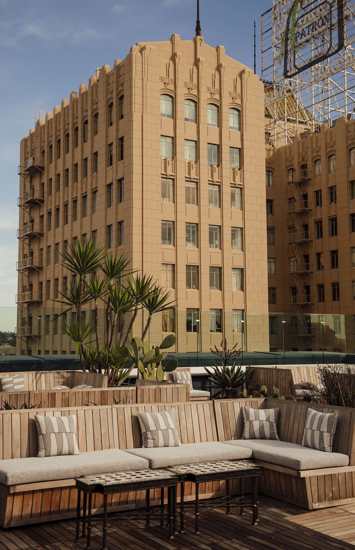 A rooftop terrace with a beige building in the background, wooden seating, and potted plants.