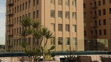 A rooftop terrace with a beige building in the background, wooden seating, and potted plants.