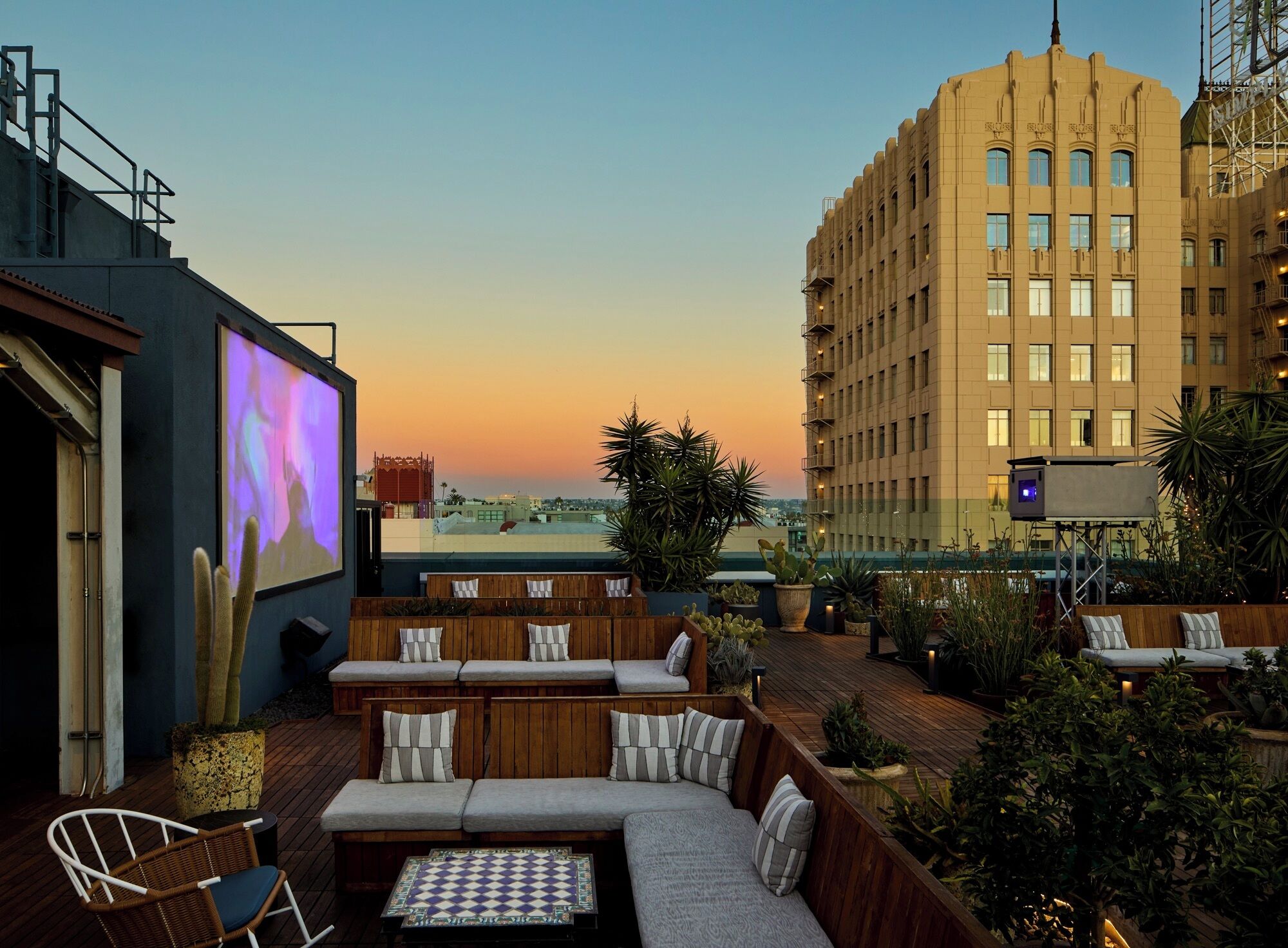 A rooftop lounge with a large screen, seating areas, and a view of a cityscape at sunset.