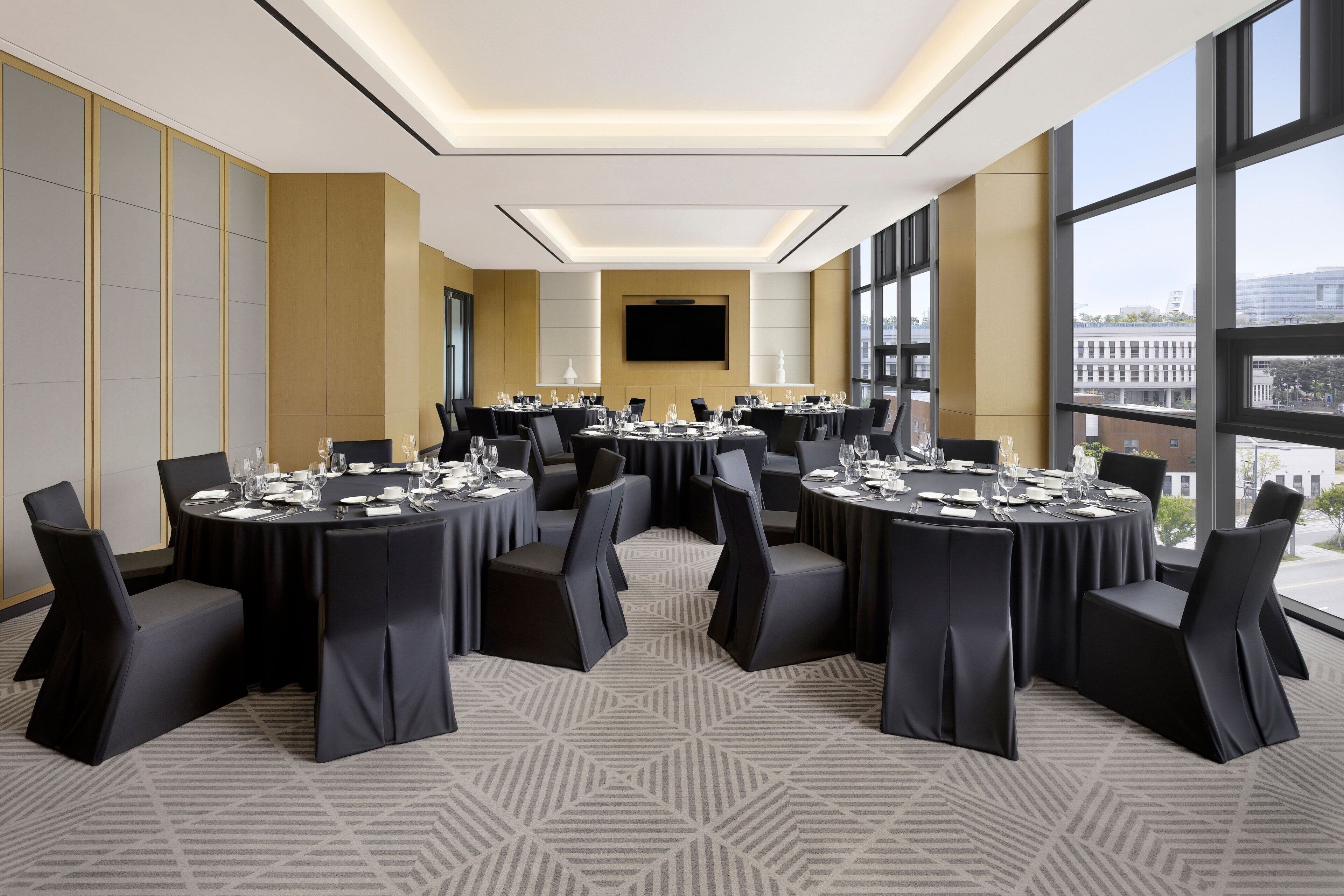 A conference room with multiple round tables, black chairs, and a large screen on the wall.