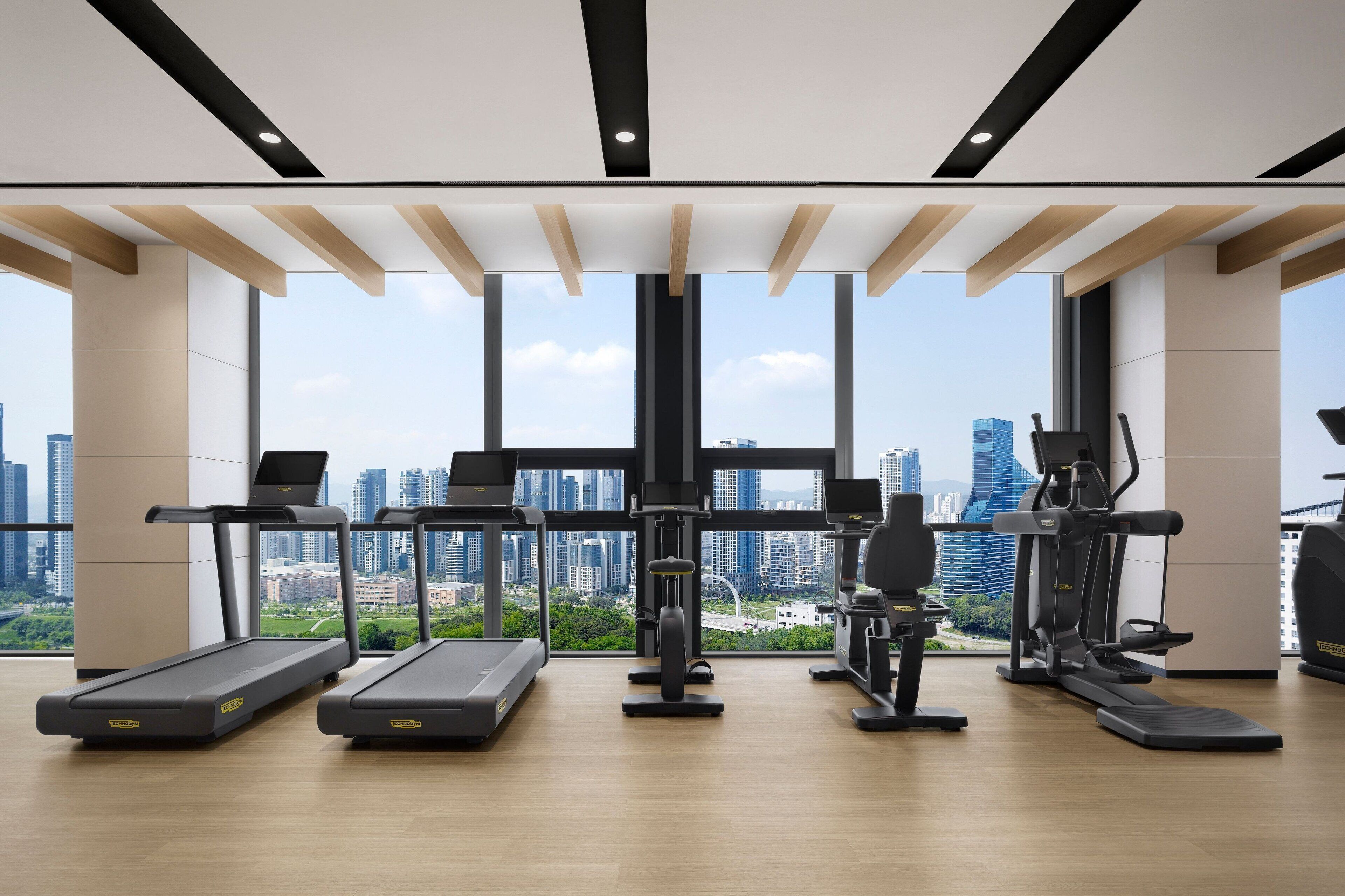 A gym with treadmills, elliptical machines, and a view of the cityscape through large windows.