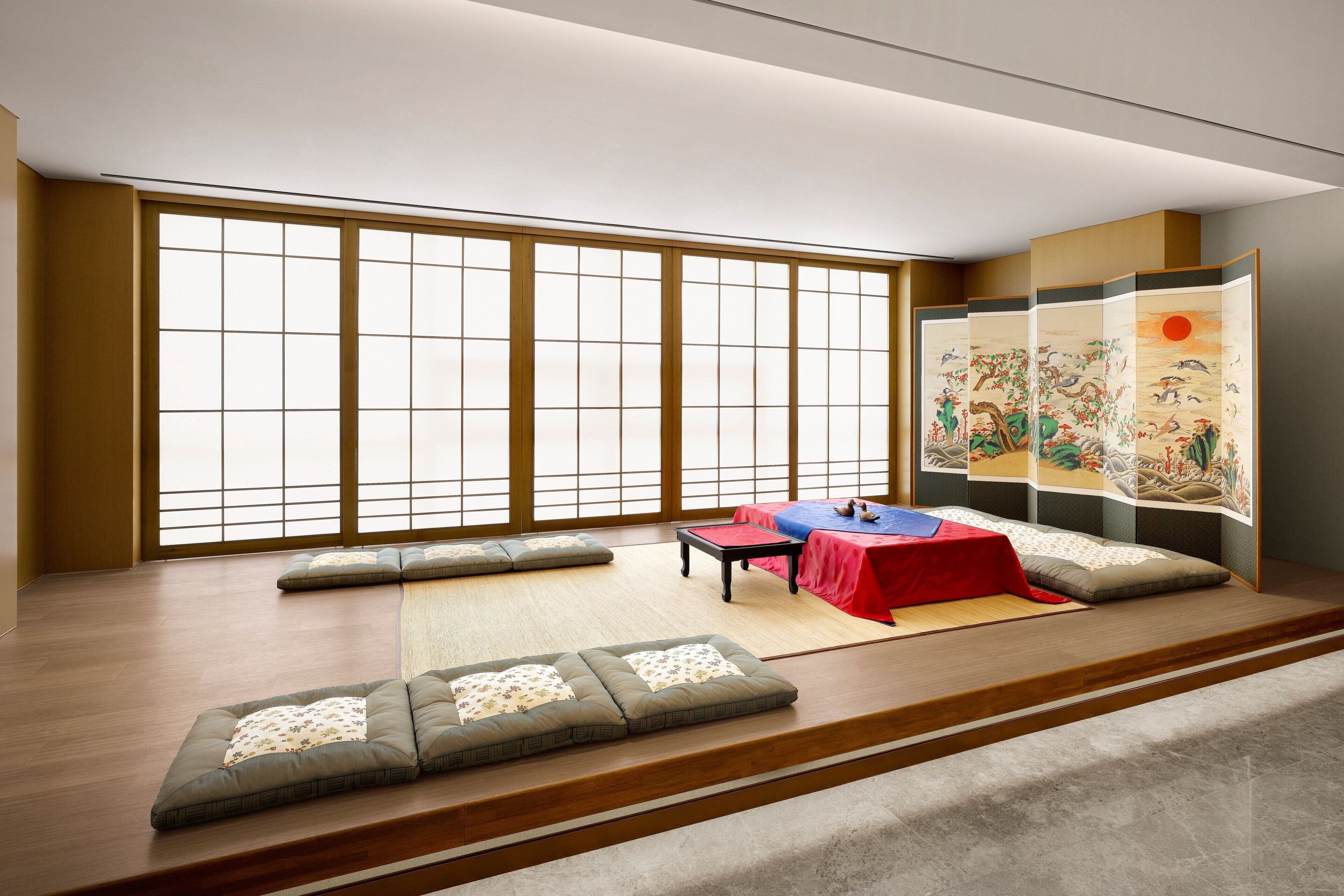 A traditional Japanese room with tatami flooring, a low table with a red tablecloth, cushions, and a sliding screen with artwork.