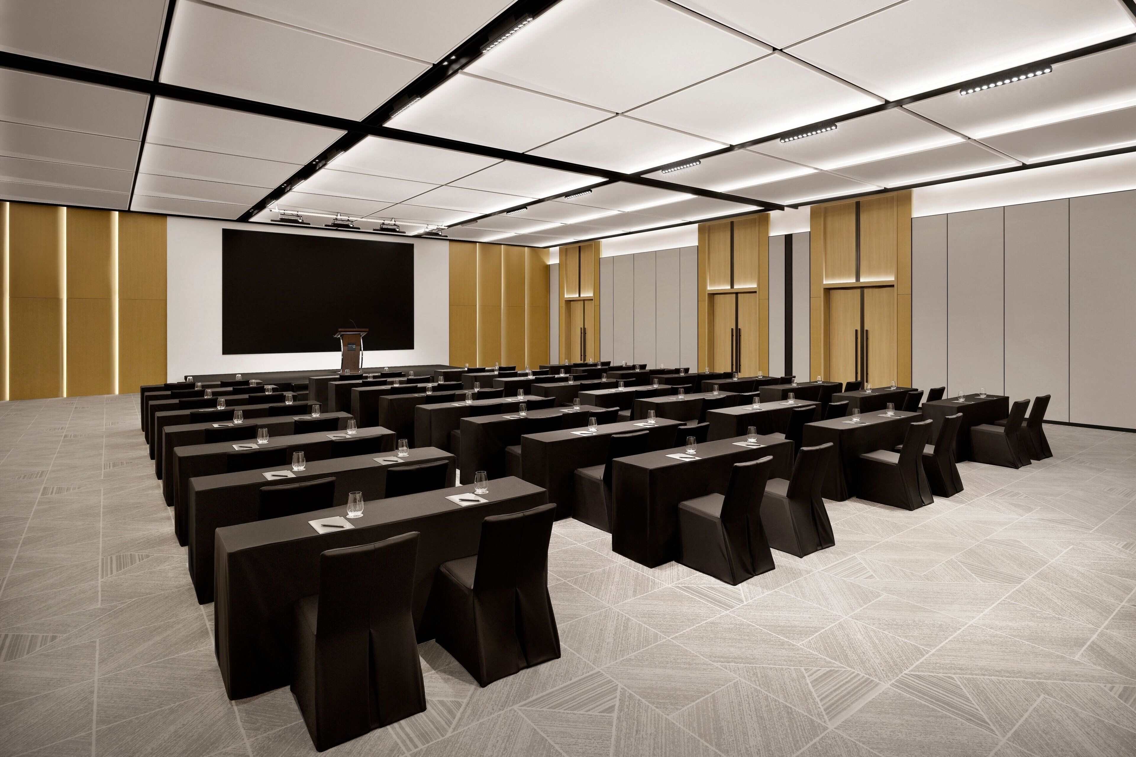 A conference room with rows of tables and chairs, a large screen, and a podium.
