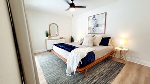 A bedroom with a bed, bedside table, nightstand, ceiling fan, and a painting on the wall.