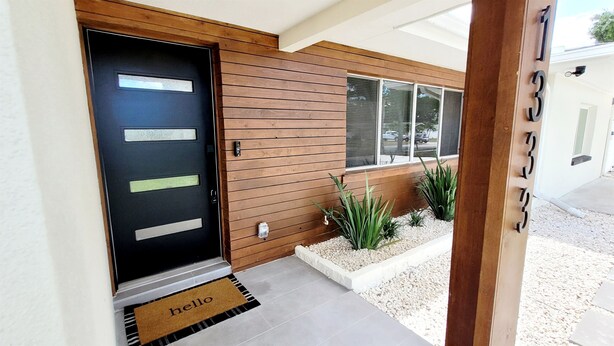 A modern house entrance with a black door, wooden siding, and a small garden area with plants and gravel.