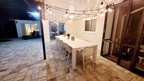 An outdoor dining area with a white table and chairs, string lights overhead, and a glass door leading to a patio.