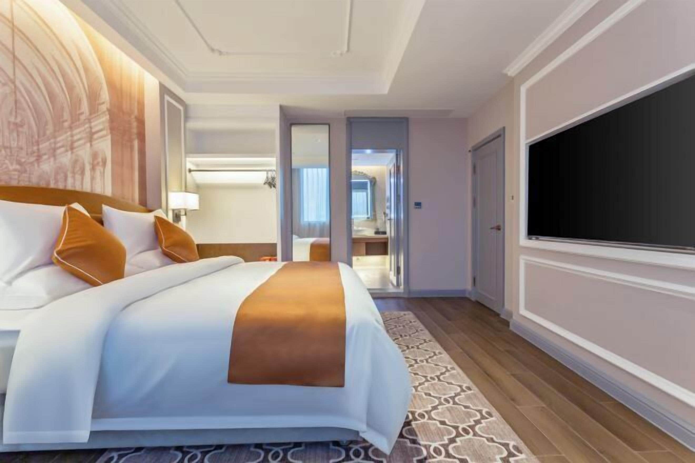 A hotel room with a large bed, a TV, a bathroom with a mirror, and a patterned rug.