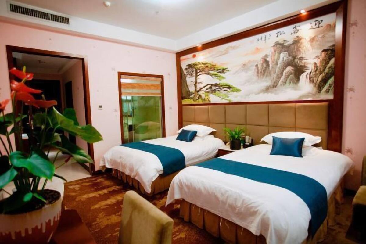 Ein Hotelzimmer mit zwei Betten, einem großen Gemälde einer Berglandschaft, einer Zimmerpflanze und einem Sessel.