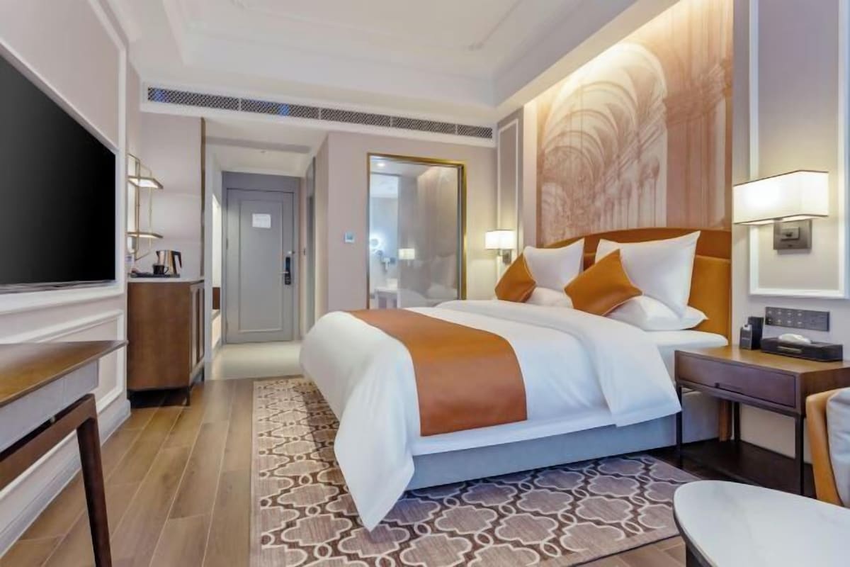 Moderni hotellihuone, jossa on suuri sänky, yöpöydät, taulutelevisio ja kuviollinen matto.