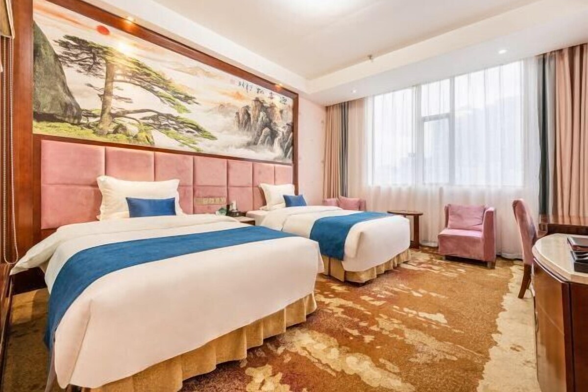 Hotellihuone, jossa on kaksi sänkyä, suuri taulu seinällä, ikkuna, jossa on verhot, ja pieni pöytä.