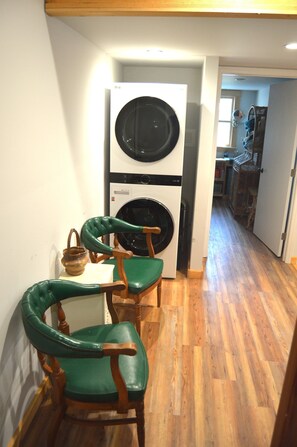 Een wasserette met een wasmachine en droger, houten stoelen en een tafeltje met een mand.
