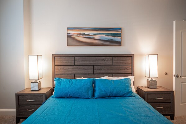 Een slaapkamer met een houten hoofdbord, een blauwgroene bedspreide, twee nachtlampjes en een schilderij van een zeegezicht.