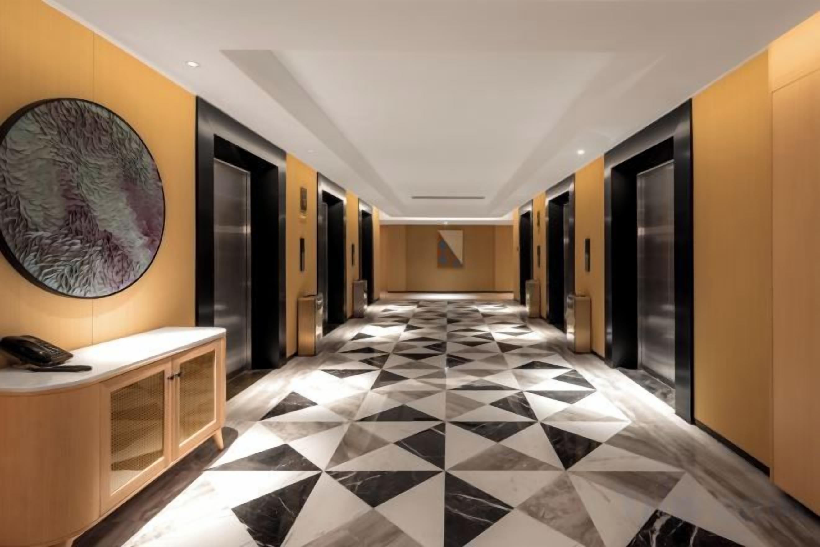 Corredor de hotel com piso de padrão geométrico, elevadores e uma grande obra de arte abstrata na parede.