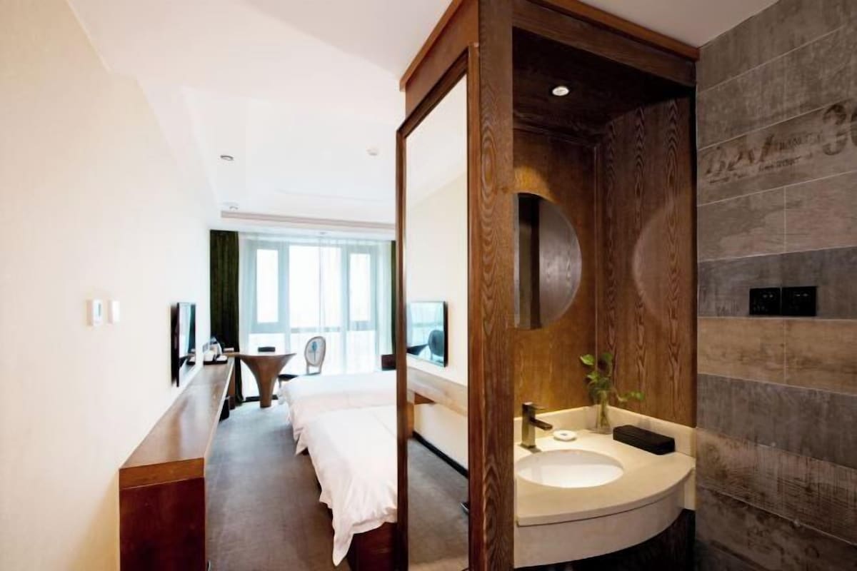 A modern bathroom with a wooden vanity, a large mirror, and a view of a bedroom with a bed and a desk.
