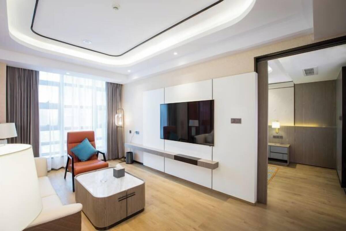 Une chambre d’hôtel moderne dotée d’un grand téléviseur à écran plat fixé au mur, d’un coin salon confortable avec un canapé et un fauteuil, ainsi que d’une table basse en bois.