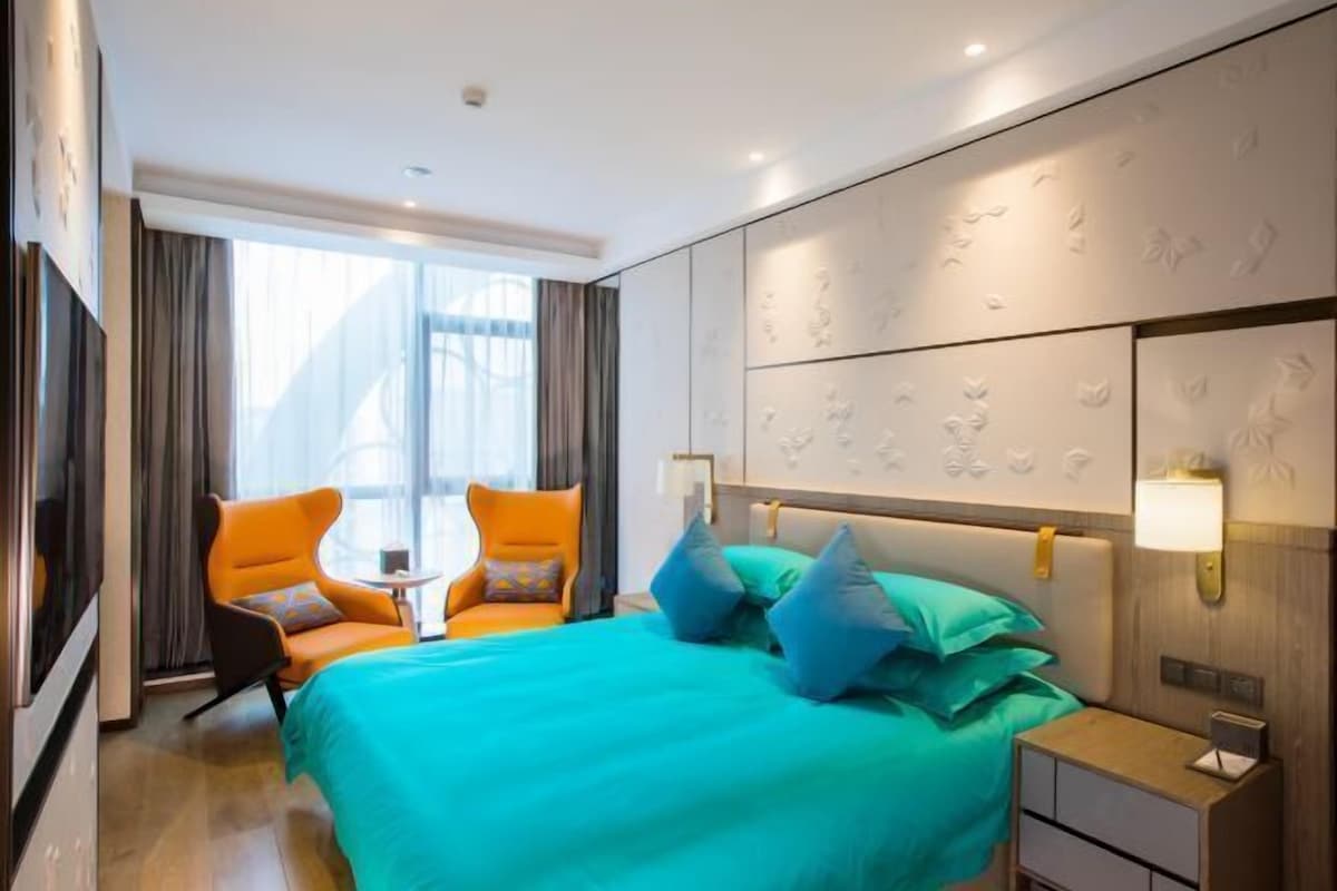 A hotel room with a large bed, two orange armchairs, a small table, and a nightstand with a lamp.
