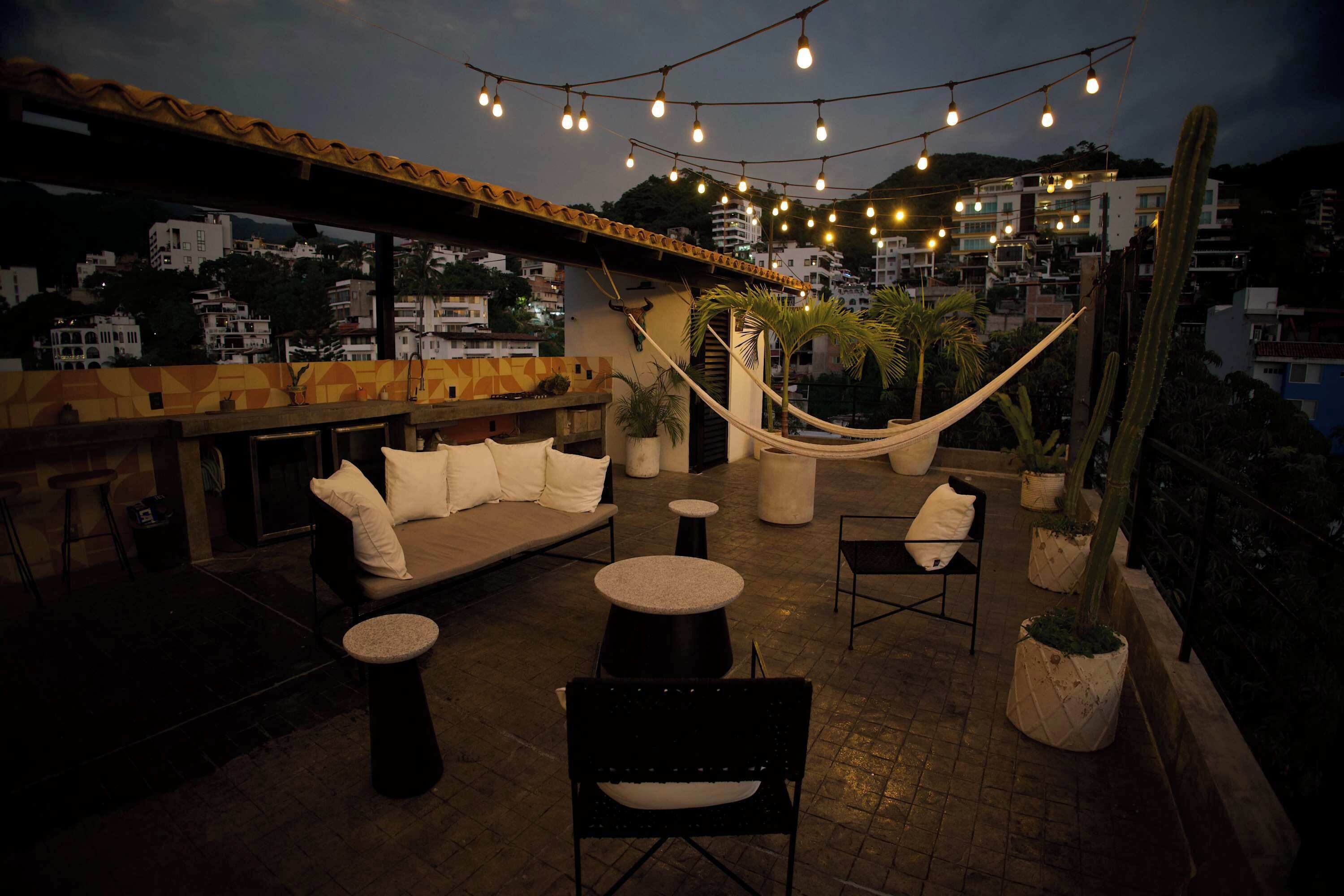 An outdoor seating area with a hammock, tables, and chairs, overlooking a cityscape.