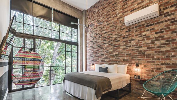 A bedroom with a brick wall, a bed, a hanging chair, and a window with a view of trees. - Loft 302 Summary:<br>We (Puerto Vallarta)