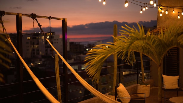 A hammock chair with a cushion, a potted plant, and string lights on a balcony with a city view at dusk. - Room 102 (Puerto Vallarta)