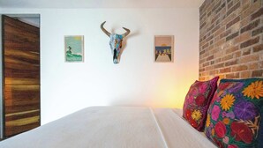 A bed with white bedding, a wooden door, a brick wall, and two framed pictures on the wall. - Room 101 (Puerto Vallarta)