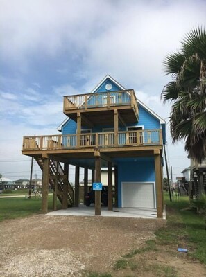Stylish two-story residence with a spacious balcony and wooden stilts, set amidst a lush green field and a palm tree.