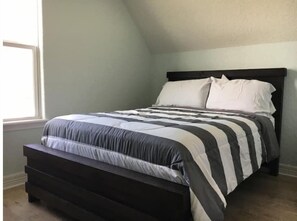 Cozy loft-style room with a central bed and dark headboard, featuring a bench at the foot and a single window.
