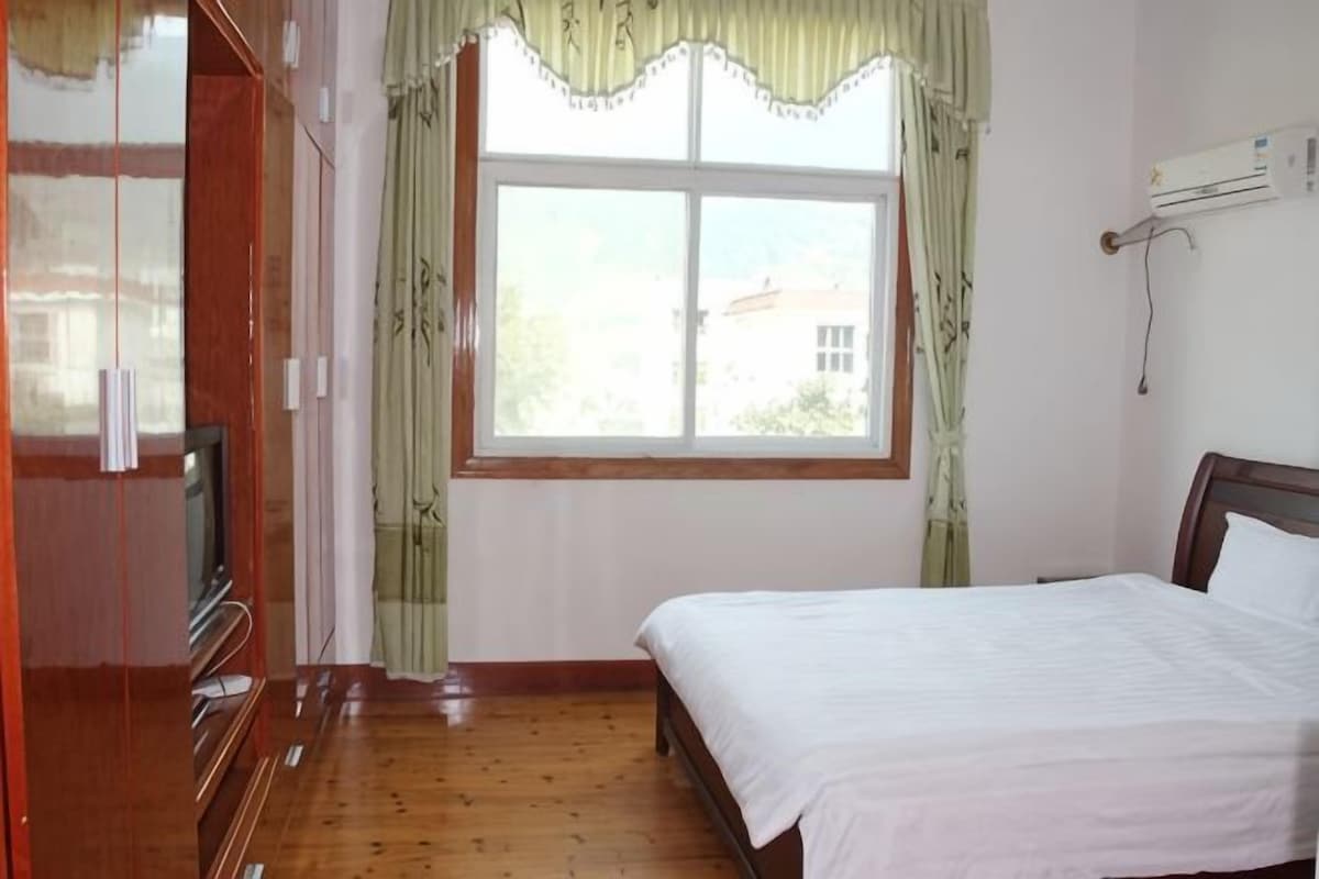 A bedroom with a bed, a wooden wardrobe, a window with curtains, and an air conditioning unit.