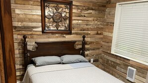 Un dormitorio con cabecera de madera, cama con colcha acolchada, un elemento decorativo en la pared sobre la cama y una ventana con persianas.