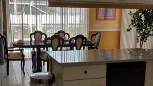 Une cuisine dotée d’un îlot central, d’un coin repas avec des chaises et une table, et offrant une vue sur l’extérieur grâce à de grandes fenêtres.