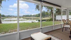 A room with large windows overlooking a lake, a grassy area, and trees. There are wicker chairs and a small table with a cushion. - Lake house retreat. (Inverness)