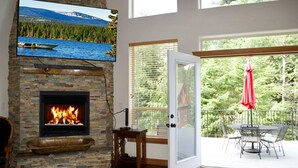 A living room with a stone fireplace, a mounted TV, and a balcony with outdoor furniture. - Beautiful home on the 10th green on Priest Lake Golf Course (Priest Lake)